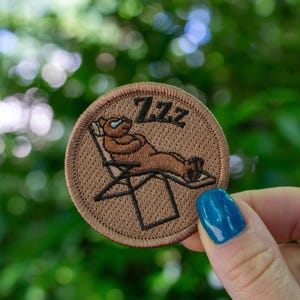 May include: A brown embroidered patch with a cartoon bear wearing sunglasses and relaxing on a lounge chair. The text "ZZZ" is above the bear.
