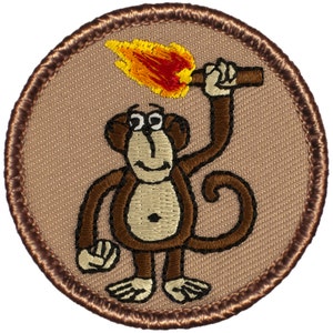 May include: A brown embroidered patch with a cartoon monkey holding a torch with a flame. The monkey is smiling and has large eyes. The patch is circular and has a brown border.