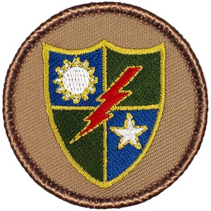 May include: A brown embroidered patch with a shield design. The shield is divided into four sections: blue, green, blue, and green. The blue sections have a white star and a white sun with rays. The green sections have a red lightning bolt. The patch is surrounded by a brown border.