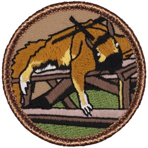 May include: A brown embroidered patch with a cartoon image of a dog lying on a wooden platform. The dog is brown with black eyes and a black nose. The platform is brown with a green background.