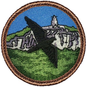 May include: A round, brown-bordered patch with an embroidered image of a black bird flying over a green hill with a white building on top. The text "The" is embroidered on the hill in purple.
