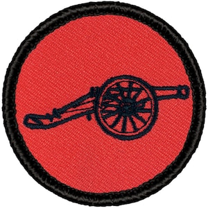 May include: A red embroidered patch with a black outline, featuring a cannon with a wheel. The cannon is facing left and has a simple design.