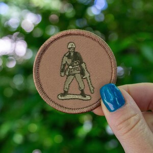May include: A brown embroidered patch with a green toy soldier holding a rifle. The patch is circular and has a brown border.