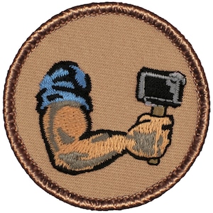 May include: A brown embroidered patch with a tan background featuring a cartoon arm holding a hammer. The arm is wearing a blue and white striped sleeve.