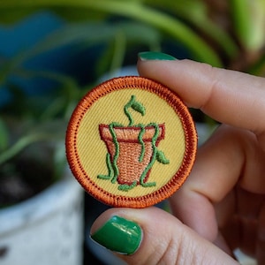 May include: A round, embroidered patch with a plant pot design. The patch features a yellow background with an orange border. The plant pot is brown, and the plant has green vines and leaves. The patch is held by a person.