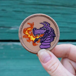 May include: A purple dragon with orange flames coming out of its mouth is embroidered on a tan patch.