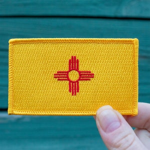 May include: A yellow embroidered patch with the state flag of New Mexico. The flag features a red Zia symbol on a yellow background.