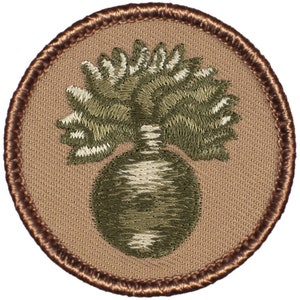 May include: A tan embroidered patch with a brown border. The patch features a green and white stylized grenade with a fuse.