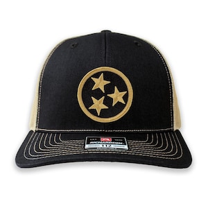 May include: Black and tan trucker hat with a gold embroidered Tennessee state flag emblem. The hat features a curved brim, mesh back, and an adjustable snap closure. The hat has the text "RICHARDSON 112 TRUCKER" on the label.