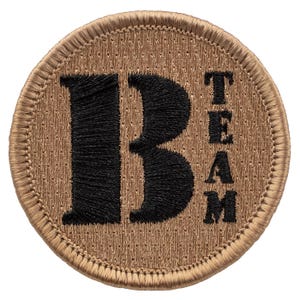 May include: A tan circular patch with black embroidered lettering. The patch says "B Team" with the "B" being large and the "Team" being smaller and vertical.