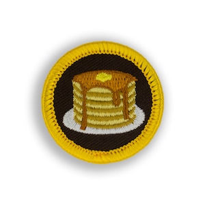 May include: A yellow embroidered patch with a brown background featuring a stack of pancakes with syrup and a pat of butter on top.