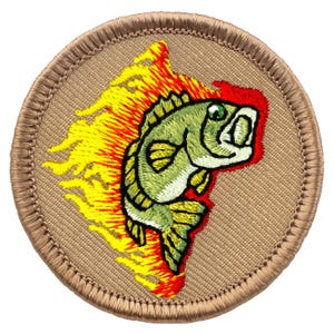 May include: Embroidered patch featuring a green fish with a wide open mouth, set against a backdrop of yellow and red flames. The fish has a single green eye. The patch is circular with a tan border and background.
