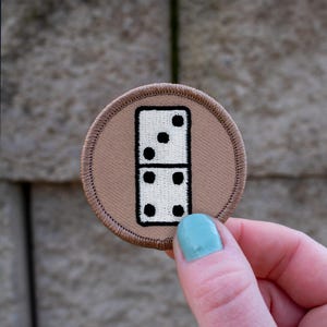May include: A brown embroidered patch with a white domino with black dots. The domino has three dots on one side and two dots on the other side.
