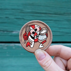 May include: A brown embroidered patch with a cartoon character of a red and white striped candy with a spoon and a bag of sugar that says "Sugar".