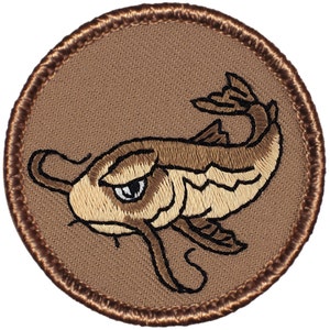 May include: Embroidered patch featuring a grumpy-looking catfish in shades of brown and tan. The fish is set against a tan background within a circular border with a brown stitched edge. Ideal for jackets, bags, or other fabric items.