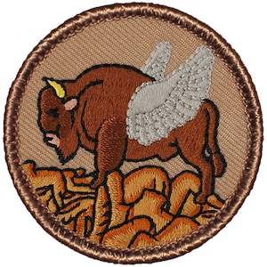May include: A brown embroidered patch with a brown bison with gray wings standing on a bed of brown grass. The patch has a brown stitched border.