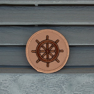 May include: A brown embroidered ship's wheel on a beige fabric patch.