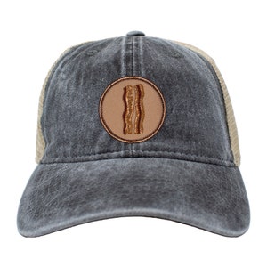 May include: A black and tan trucker hat with a brown circle patch featuring a cartoon image of two strips of bacon.