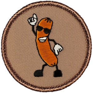 May include: A brown embroidered patch with a cartoon sausage wearing sunglasses and a smile. The sausage is holding its arm up in the air and has a black and white outline.