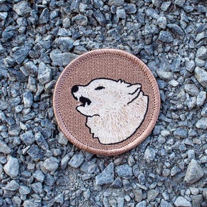 May include: A brown embroidered patch with a white wolf howling at the moon. The wolf is outlined in black thread.