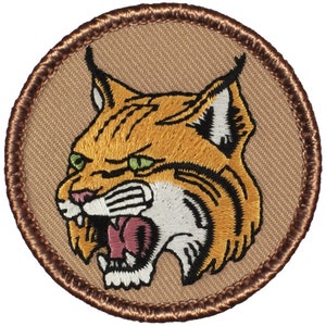 May include: A brown embroidered patch with a tan background featuring a snarling bobcat with green eyes. The patch has a brown stitched border.