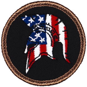 May include: A round, brown embroidered patch with a black background. The patch features a white and red American flag design in the shape of a Spartan helmet.