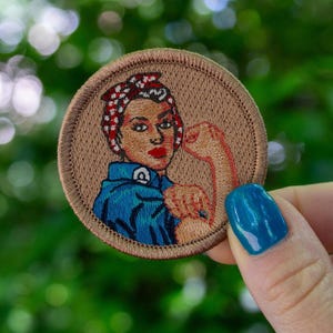 May include: A brown embroidered patch with a woman wearing a red, white, and blue bandana and a blue shirt with a raised fist. The text "We Can Do It" is embroidered on the shirt.