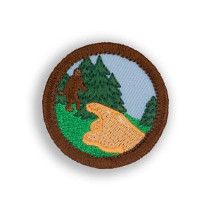 May include: A brown embroidered patch with a green background featuring a brown Bigfoot figure walking in a forest with a hand pointing to the right. The patch has a brown border.
