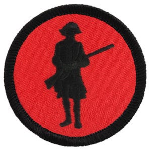 May include: A circular embroidered patch featuring a black silhouette of a person in 18th-century attire holding a long rifle. The design is set against a vibrant red background and is finished with a thick, black stitched border around the edge.
