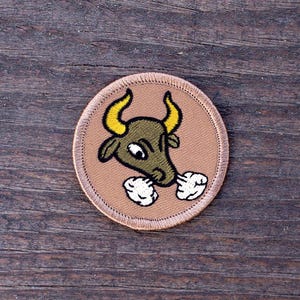 May include: A brown embroidered patch with a cartoon bull with yellow horns and white puffs of air coming from its nostrils.