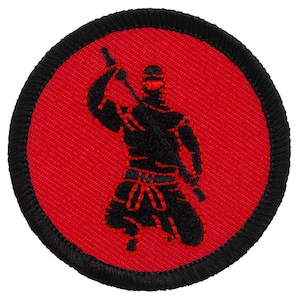 May include: A circular embroidered patch featuring a vibrant red background with a black border. The center displays a black silhouette of a kneeling ninja figure holding a sword above its head, with red stitching details accentuating the clothing.
