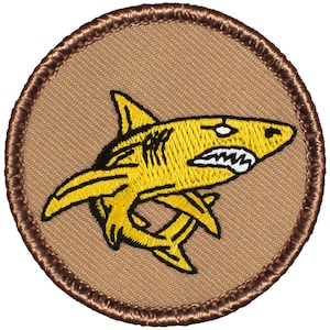 May include: A yellow embroidered shark patch with a brown stitched border. The shark is facing right and has a white eye and teeth.