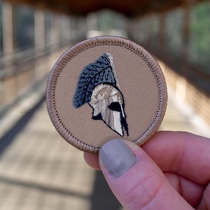 May include: A brown embroidered patch with a white and black Spartan helmet design.