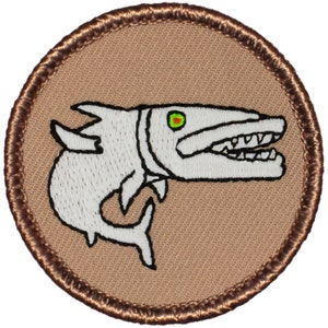 May include: A brown embroidered patch with a white cartoon fish with a green eye and open mouth. The fish is on a tan background.