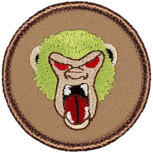 May include: A round embroidered patch with a brown border. The center of the patch features a green, snarling monkey face with red eyes and a red tongue.