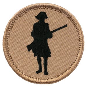 May include: A circular embroidered patch featuring a tan background with a dark brown stitched border. In the center, a black silhouette of a standing figure wearing a tricorn hat and a long coat, holding a long rifle across the body.