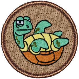 May include: A brown embroidered patch with a green turtle with a yellow shell and brown eyes. The turtle is in a relaxed pose with its head tilted to the side. The patch has a brown border.