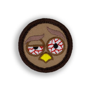 May include: A brown embroidered patch with a cartoon face with red bloodshot eyes and a yellow beak.