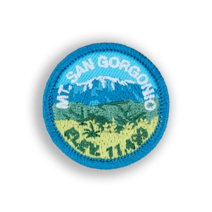 May include: A round embroidered patch with a blue border. The patch features a mountain scene with the text "MT. SAN GORGONIO" and "RE2 11 APR". The design includes blue, green, and yellow colors.