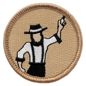 May include: A brown embroidered patch with a white outline. The patch features a cartoon figure of a person wearing a black and white striped shirt, black pants, a black hat with a white stripe, and a white beard. The person is holding their right arm up in the air with their index finger pointing up.