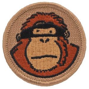 May include: Embroidered patch featuring a brown and orange cartoon monkey face with a black headband. The patch is circular and has a tan border.