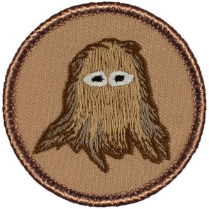 May include: A brown embroidered patch with a cartoon ghost with white eyes. The patch is circular and has a brown stitched border.