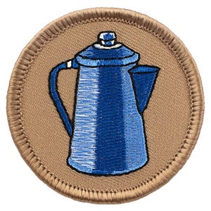 May include: A circular embroidered patch featuring a blue coffee percolator with a white lid knob and handle detail, set against a tan background with a thick, stitched tan border. The design uses shades of blue thread to create depth.