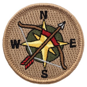 May include: A tan embroidered patch with a compass rose and two crossed arrows. The compass rose is yellow with a green background. The arrows are brown with red fletching. The patch has a tan border with a stitched edge.