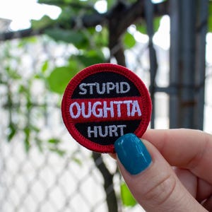 May include: A round, embroidered patch with the words "STUPID OUGHTTA HURT" in white lettering. The patch has a black background with a red border and a horizontal red stripe. The patch is held by a hand with blue nail polish.