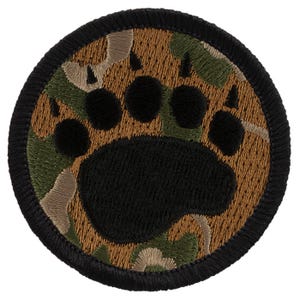 May include: A round, black embroidered patch with a brown and green camouflage pattern. The patch features a black bear paw print in the center.