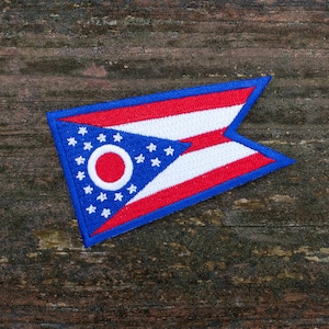 May include: Embroidered patch featuring the flag of Ohio. The patch is shaped like a pennant, with a blue triangle containing a white circle and stars. Red and white stripes complete the design, with a blue border.