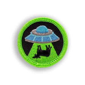 May include: Round embroidered patch featuring a blue and silver UFO beaming up a black cow with a green beam. The patch has a black background and a bright green border.