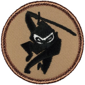 May include: A black and white embroidered patch of a ninja holding a sword. The patch is circular and has a brown border.
