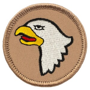 May include: A tan embroidered patch with a white eagle head with a yellow beak and red mouth. The eagle is facing left.